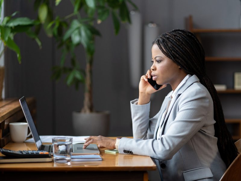 afro-female-business-consultant-interacting-with-client-over-phone-and-entering-data-on-laptop