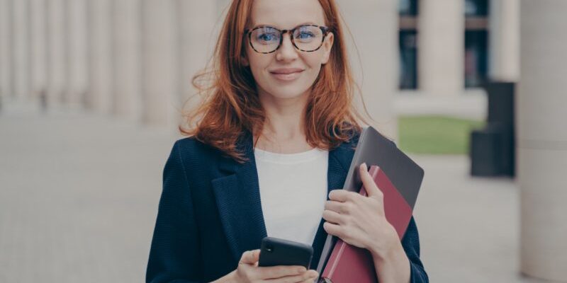 confident-beautiful-red-haired-female-business-consultant-holding-modern-smartphone-and-laptop-800x533